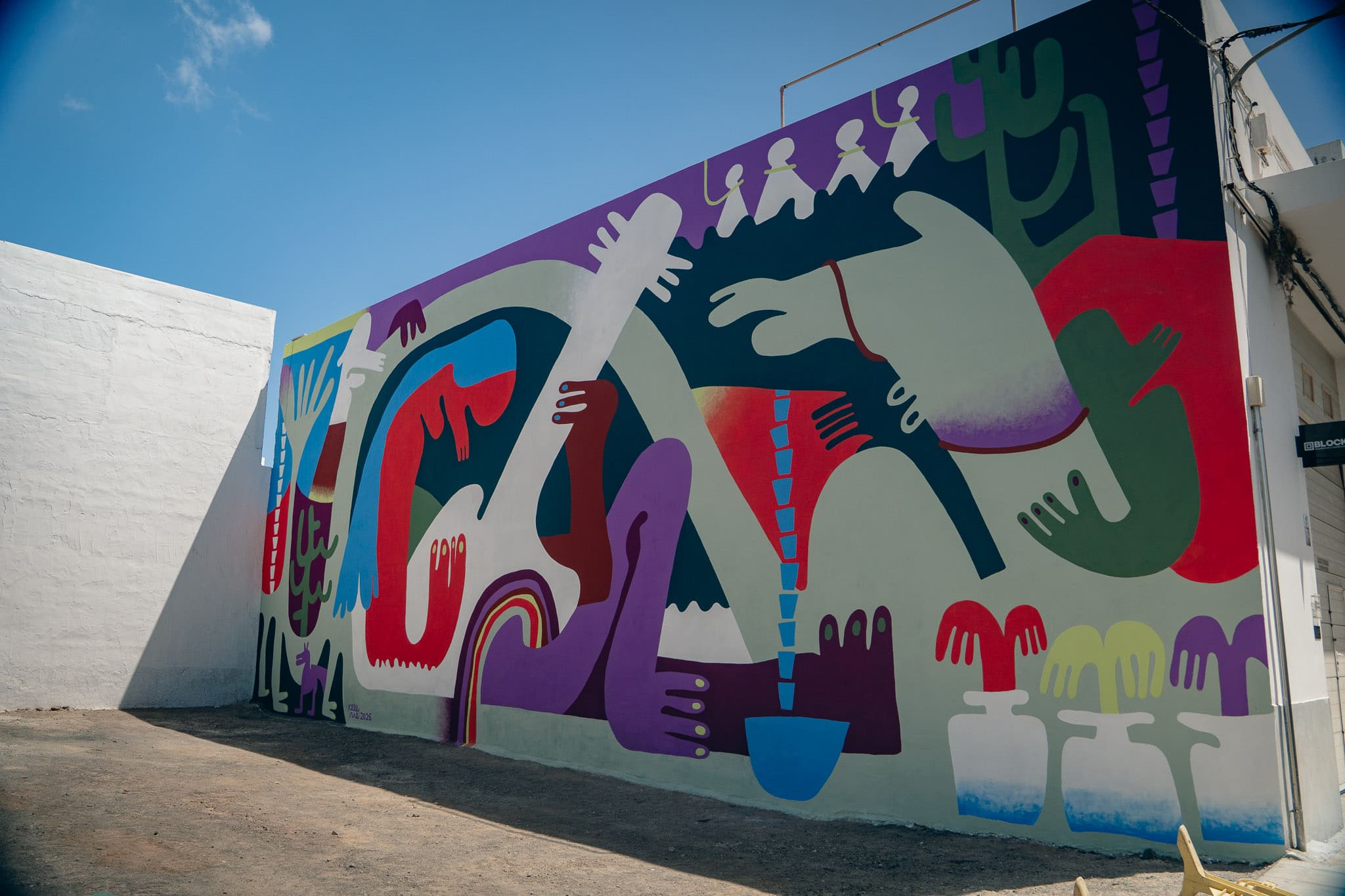 EL MURO QUE NO SE REFLEJA EN EL ALJIBE Antonia Brito «No te alongues al aljibe», nos gritaban de chinijos. La advertencia formaba parte de la vida cotidiana de Lanzarote y nos prevenía del peligro de asomarnos a aquella profundidad líquida y oscura. Hoy nos recuerda también a una isla que parece olvidar el papel que sus aljibes desempeñaron —y todavía deberían desempeñar—, obligada durante siglos a guindar y aprovechar cada gota de agua. Los artistas de la tercera edición del Festival Desidia – III Circuito de Murales de Argana Baja tampoco quisieron alongarse. Aunque el lema elegido era «El aljibe. Memoria subterránea», apenas se acercaron a su historia, a su significado patrimonial o a la grave escasez de agua que padece Lanzarote. El título, cargado de evocaciones, quedó suspendido sobre las obras como una etiqueta que nadie mira. Prometía profundidad, pero lo que debía constituir el corazón del proyecto acabó reducido, en buena medida, a su envoltorio. El problema no es que el arte hable de identidad, disidencia o experiencias personales. Todos ellos son asuntos legítimos y necesarios. El problema es anunciar el aljibe lanzaroteño como eje del festival para vaciarlo después de su historia material y reducirlo a una metáfora tan amplia que termine justificando prácticamente cualquier obra. Tampoco el programa dedicó una actividad específica a los aljibes, las maretas o los antiguos sistemas de captación de la lluvia. No hubo, al menos en la programación presentada al público, una aproximación histórica, patrimonial o medioambiental que ayudara a comprender por qué esas construcciones resultaron esenciales para la supervivencia de Lanzarote. Pero los aljibes no pertenecen solamente al pasado. La dependencia del petróleo para producir agua y la creciente exposición de Canarias a fenómenos ciclónicos podrían obligarnos, en el futuro, a volver a necesitarlos. LA INTERNACIONALIDAD LLEGÓ ANTES QUE LA DOCUMENTACIÓN La presencia de artistas procedentes de distintos lugares podía aportar otras miradas sobre Lanzarote. Sin embargo, para trabajar con un asunto tan estrechamente vinculado a la historia y al territorio era necesario ofrecerles previamente información, documentación y contacto con esa realidad. Durante la visita, una artista reconoció que, antes de llegar, imaginaba Lanzarote como una isla verde. Otra habló de los aljibes como si fueran necesariamente construcciones redondas. Se mencionó incluso una búsqueda rápida en internet para tratar de averiguar qué era aquello sobre lo que se les había pedido trabajar. La internacionalidad llegó antes que la documentación. Los artistas aterrizaron en una isla cuya memoria debían interpretar sin haber tenido tiempo ni herramientas suficientes para conocerla. El resultado no puede atribuirse únicamente a quienes pintaron los muros. También revela una carencia en la concepción y preparación del proyecto. Otro artista partió de una cortina vinculada a su memoria familiar para hablar de cambios, rupturas y de un proceso identitario personal. Se trata de un relato legítimo por sí mismo, pero su relación con el aljibe quedó débilmente explicada. Una cortina puede contener memoria familiar, pero el aljibe contenía la memoria colectiva de toda una isla. Reducir un asunto histórico, patrimonial y medioambiental de semejante densidad a asociaciones personales resulta una aproximación demasiado superficial, casi mezquina frente a la profundidad que ofrecía el tema. Las referencias familiares y los procesos individuales fueron ocupando el espacio que debía corresponder a una reflexión sobre el agua, el territorio y el patrimonio compartido. Las obras parecían surgir de universos creativos anteriores al festival, mientras la relación con el lema se añadía después mediante explicaciones. LA CURADORA ACHICA EL VACÍO Ante aquella desconexión, la curadora Elena García asumió la tarea de hilar las obras y vincularlas con la temática común. Tuvo que llenar con palabras lo que los murales habían dejado seco. Su discurso hablaba de memoria, territorio, agua y raíces. Los artistas, en cambio, describían búsquedas rápidas, referencias familiares y universos creativos que parecían existir antes de conocer el título del festival. Por momentos, la curadora encontraba en las obras una profundidad que sus propios autores no parecían reconocer. Más que revelar el camino creativo de los muralistas, tendía después los puentes necesarios para que todos llegaran, aunque fuera verbalmente, hasta el aljibe. Una obra no tiene que representar literalmente una cisterna ni convertirse en una lección ilustrada de patrimonio. El arte puede ser abstracto, simbólico, íntimo y abierto. Pero si un festival adopta como eje «El aljibe. Memoria subterránea», sería razonable que ofreciera a los participantes algo más que un muro, pintura y una conexión a internet. Cuando un proyecto sostenido con recursos públicos adopta una temática patrimonial para proporcionar sentido al conjunto, cabe exigir seriedad y compromiso con aquello que anuncia. El tema debe penetrar en la documentación, en la investigación y en el proceso creativo; no limitarse a decorar posteriormente un discurso. El aljibe funcionó como reclamo y como promesa de profundidad ante una sociedad preocupada por el abandono de su patrimonio. Sin embargo, apenas penetró en la investigación, en el programa o en las obras. La temática tuvo que ser reconstruida después mediante el discurso curatorial. UNA OPORTUNIDAD PERDIDA El festival podía haber mostrado que recuperar los aljibes no es solo conservar un bien histórico, sino prepararnos para el futuro. Lanzarote depende de una desalación sostenida, en gran medida, por combustibles fósiles, mientras el calentamiento del Atlántico acerca a Canarias fenómenos ciclónicos hasta ahora excepcionales. Ante una crisis energética, una avería o una catástrofe, recoger y almacenar el agua de lluvia constituye una reserva esencial y una forma de reducir nuestra vulnerabilidad. También podía haber acercado a los artistas y a la ciudadanía a los sistemas tradicionales de captación de agua, al trabajo de quienes cuidaban los aljibes y al abandono de muchos de ellos. Había agua suficiente para una reflexión artística, histórica, social y medioambiental profunda. Sin embargo, casi nadie quiso alongarse. Las trayectorias y los conflictos personales ocuparon el lugar que correspondía a la memoria colectiva. La temática tuvo que ser reconstruida después mediante el discurso curatorial. No faltaba tema. Faltó profundidad. El resultado es un título hermoso, pero en buena medida vacío: tan vacío como muchos de los aljibes abandonados que el festival invoca sin llegar realmente a mirar. EL MURO QUE NO SE REFLEJA EN EL ALJIBE Antonia Brito «No te alongues al aljibe», nos gritaban de chinijos. La advertencia formaba parte de la vida cotidiana de Lanzarote y nos prevenía del peligro de asomarnos a aquella profundidad líquida y oscura. Hoy nos recuerda también a una isla que parece olvidar el papel que sus aljibes desempeñaron —y todavía deberían desempeñar—, obligada durante siglos a guindar y aprovechar cada gota de agua. Los artistas de la tercera edición del Festival Desidia – III Circuito de Murales de Argana Baja tampoco quisieron alongarse. Aunque el lema elegido era «El aljibe. Memoria subterránea», apenas se acercaron a su historia, a su significado patrimonial o a la grave escasez de agua que padece Lanzarote. El título, cargado de evocaciones, quedó suspendido sobre las obras como una etiqueta que nadie mira. Prometía profundidad, pero lo que debía constituir el corazón del proyecto acabó reducido, en buena medida, a su envoltorio. El problema no es que el arte hable de identidad, disidencia o experiencias personales. Todos ellos son asuntos legítimos y necesarios. El problema es anunciar el aljibe lanzaroteño como eje del festival para vaciarlo después de su historia material y reducirlo a una metáfora tan amplia que termine justificando prácticamente cualquier obra. Tampoco el programa dedicó una actividad específica a los aljibes, las maretas o los antiguos sistemas de captación de la lluvia. No hubo, al menos en la programación presentada al público, una aproximación histórica, patrimonial o medioambiental que ayudara a comprender por qué esas construcciones resultaron esenciales para la supervivencia de Lanzarote. Pero los aljibes no pertenecen solamente al pasado. La dependencia del petróleo para producir agua y la creciente exposición de Canarias a fenómenos ciclónicos podrían obligarnos, en el futuro, a volver a necesitarlos. LA INTERNACIONALIDAD LLEGÓ ANTES QUE LA DOCUMENTACIÓN La presencia de artistas procedentes de distintos lugares podía aportar otras miradas sobre Lanzarote. Sin embargo, para trabajar con un asunto tan estrechamente vinculado a la historia y al territorio era necesario ofrecerles previamente información, documentación y contacto con esa realidad. Durante la visita, una artista reconoció que, antes de llegar, imaginaba Lanzarote como una isla verde. Otra habló de los aljibes como si fueran necesariamente construcciones redondas. Se mencionó incluso una búsqueda rápida en internet para tratar de averiguar qué era aquello sobre lo que se les había pedido trabajar. La internacionalidad llegó antes que la documentación. Los artistas aterrizaron en una isla cuya memoria debían interpretar sin haber tenido tiempo ni herramientas suficientes para conocerla. El resultado no puede atribuirse únicamente a quienes pintaron los muros. También revela una carencia en la concepción y preparación del proyecto. Otro artista partió de una cortina vinculada a su memoria familiar para hablar de cambios, rupturas y de un proceso identitario personal. Se trata de un relato legítimo por sí mismo, pero su relación con el aljibe quedó débilmente explicada. Una cortina puede contener memoria familiar, pero el aljibe contenía la memoria colectiva de toda una isla. Reducir un asunto histórico, patrimonial y medioambiental de semejante densidad a asociaciones personales resulta una aproximación demasiado superficial, casi mezquina frente a la profundidad que ofrecía el tema. Las referencias familiares y los procesos individuales fueron ocupando el espacio que debía corresponder a una reflexión sobre el agua, el territorio y el patrimonio compartido. Las obras parecían surgir de universos creativos anteriores al festival, mientras la relación con el lema se añadía después mediante explicaciones. LA CURADORA ACHICA EL VACÍO Ante aquella desconexión, la curadora Elena García asumió la tarea de hilar las obras y vincularlas con la temática común. Tuvo que llenar con palabras lo que los murales habían dejado seco. Su discurso hablaba de memoria, territorio, agua y raíces. Los artistas, en cambio, describían búsquedas rápidas, referencias familiares y universos creativos que parecían existir antes de conocer el título del festival. Por momentos, la curadora encontraba en las obras una profundidad que sus propios autores no parecían reconocer. Más que revelar el camino creativo de los muralistas, tendía después los puentes necesarios para que todos llegaran, aunque fuera verbalmente, hasta el aljibe. Una obra no tiene que representar literalmente una cisterna ni convertirse en una lección ilustrada de patrimonio. El arte puede ser abstracto, simbólico, íntimo y abierto. Pero si un festival adopta como eje «El aljibe. Memoria subterránea», sería razonable que ofreciera a los participantes algo más que un muro, pintura y una conexión a internet. Cuando un proyecto sostenido con recursos públicos adopta una temática patrimonial para proporcionar sentido al conjunto, cabe exigir seriedad y compromiso con aquello que anuncia. El tema debe penetrar en la documentación, en la investigación y en el proceso creativo; no limitarse a decorar posteriormente un discurso. El aljibe funcionó como reclamo y como promesa de profundidad ante una sociedad preocupada por el abandono de su patrimonio. Sin embargo, apenas penetró en la investigación, en el programa o en las obras. La temática tuvo que ser reconstruida después mediante el discurso curatorial. UNA OPORTUNIDAD PERDIDA El festival podía haber mostrado que recuperar los aljibes no es solo conservar un bien histórico, sino prepararnos para el futuro. Lanzarote depende de una desalación sostenida, en gran medida, por combustibles fósiles, mientras el calentamiento del Atlántico acerca a Canarias fenómenos ciclónicos hasta ahora excepcionales. Ante una crisis energética, una avería o una catástrofe, recoger y almacenar el agua de lluvia constituye una reserva esencial y una forma de reducir nuestra vulnerabilidad. También podía haber acercado a los artistas y a la ciudadanía a los sistemas tradicionales de captación de agua, al trabajo de quienes cuidaban los aljibes y al abandono de muchos de ellos. Había agua suficiente para una reflexión artística, histórica, social y medioambiental profunda. Sin embargo, casi nadie quiso alongarse. Las trayectorias y los conflictos personales ocuparon el lugar que correspondía a la memoria colectiva. La temática tuvo que ser reconstruida después mediante el discurso curatorial. No faltaba tema. Faltó profundidad. El resultado es un título hermoso, pero en buena medida vacío: tan vacío como muchos de los aljibes abandonados que el festival invoca sin llegar realmente a mirar.